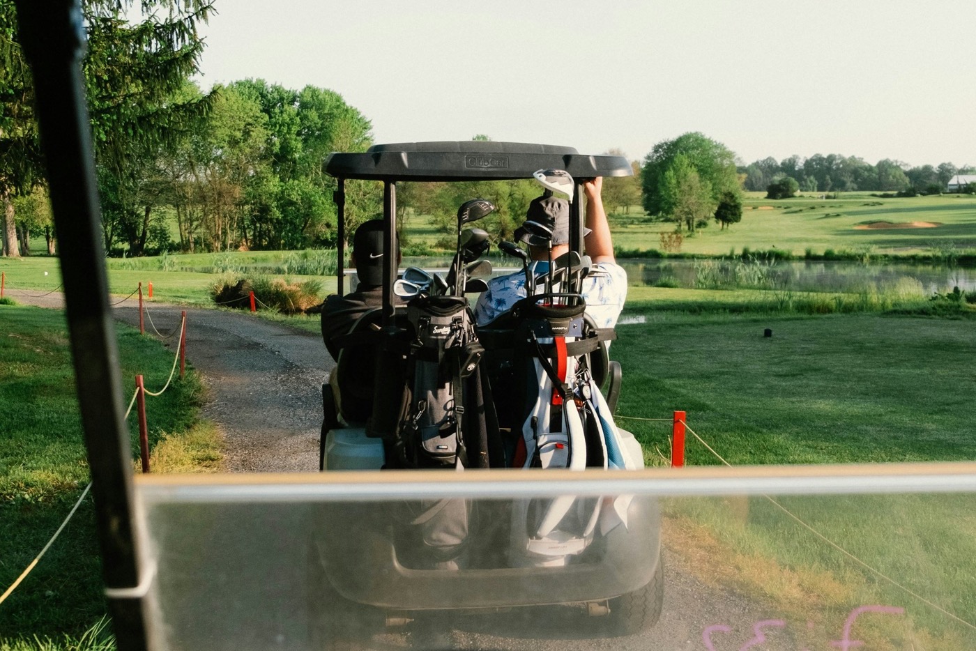 Two golfers riding together in a golf cart on a sunny course, seen from a following cart.